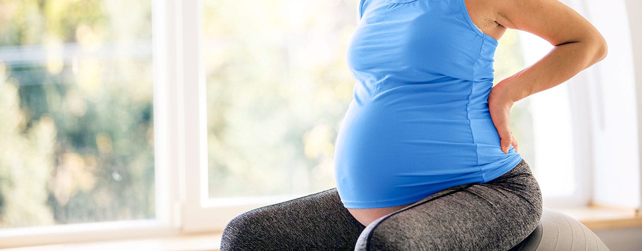 Mid adult pregnant woman is holding her arms on her back, feeling a bit pain and sitting on a fitness ball at home