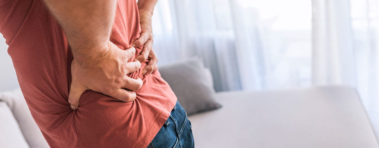 Photo of unhappy man suffering from backache at home while standing during the day. Close up of mature man rubbing his painful back. Pain relief, chiropractic concept